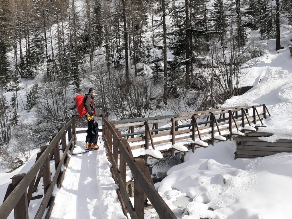 Ludovic, le co-fondateur d'Outdoor Fix, le service d'entretien de matériel outdoor, est en train de traverser à ski de randonnée un pont enneigé sur une rivière dans une forêt.