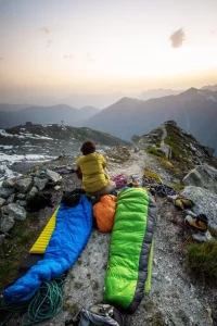 expédition en montagne, pause avec les duvets étalés sur le sol, une personne assise, de dos, regarde les montagnes à l'horizon